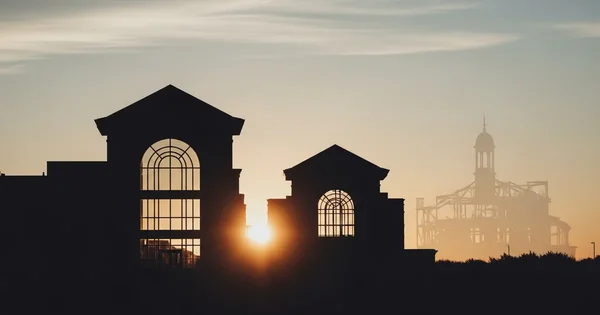 The sun rises behind silhouetted buildings and construction in an unidentifiable location.