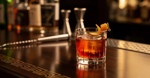A craft cocktail garnished with an orange twist sits on a polished bar top alongside professional mixing tools.