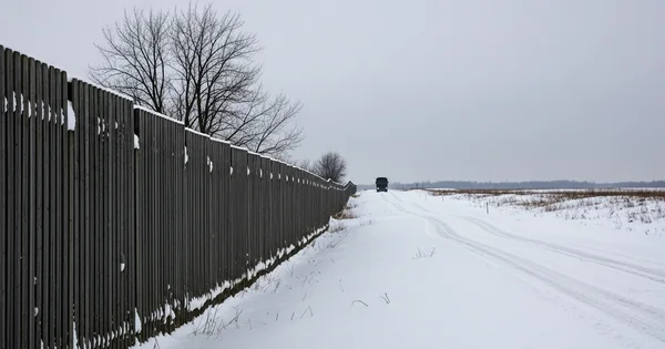 A vehicle travels along a snow-covered road beside a long wooden fence.
