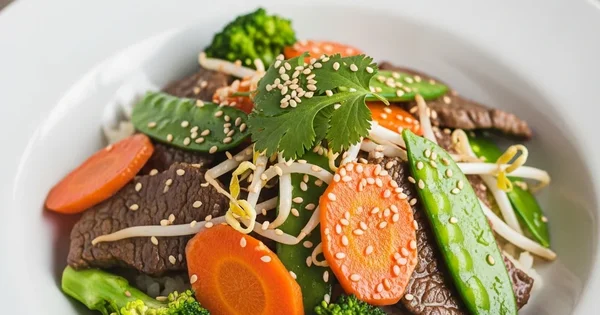 A bowl of beef stir fry with carrots, broccoli, and snow peas is topped with sesame seeds and cilantro.