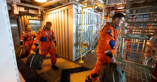 Astronauts in orange spacesuits walk through a facility carrying bags.