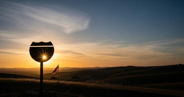 The sun sets behind a roadside memorial sign and an American flag overlooking a winding highway through rolling hills.
