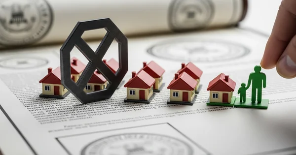 Miniature houses sit on a document with a hand placing a family figure nearby.