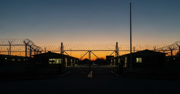 A gate with barbed wire stands in front of a prison at dusk.